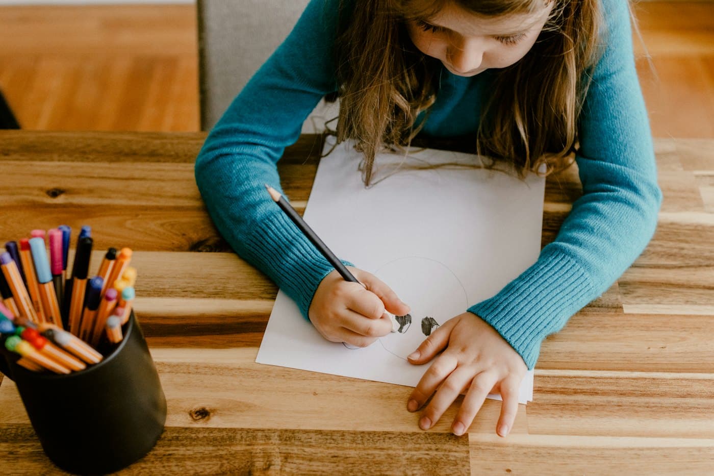 A child drawing on paper at a wooden table, with a cup of colored pencils beside them.